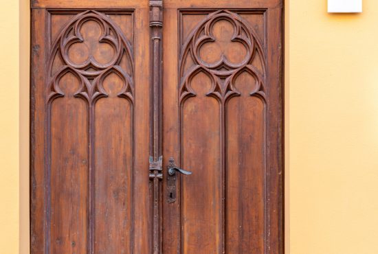 Tall wooden doors with gothic carvings stand in a yellow, orange wall marked with the number thirteen. Warm tones and grainy timber texture give the entrance a strong historic character.